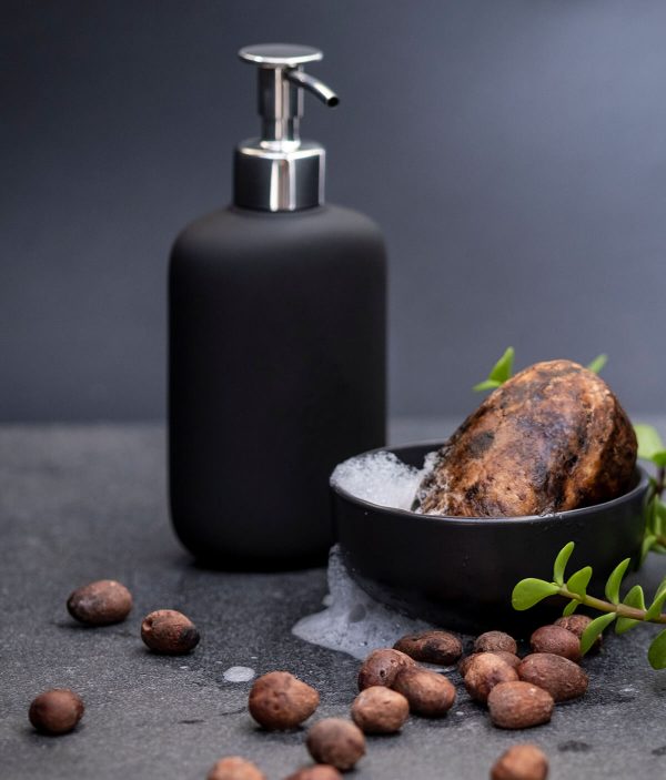 Deluxe African Black Soap bar with suds in a black bowl next to a pump bottle, with Shea nuts scattered decoratively on the floor.