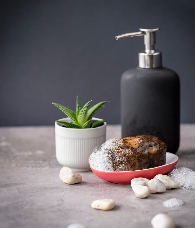 Deluxe African Black Soap bar with suds in a red dish next to a pump bottle and small pot plant, with pebbles scattered decoratively on the floor.