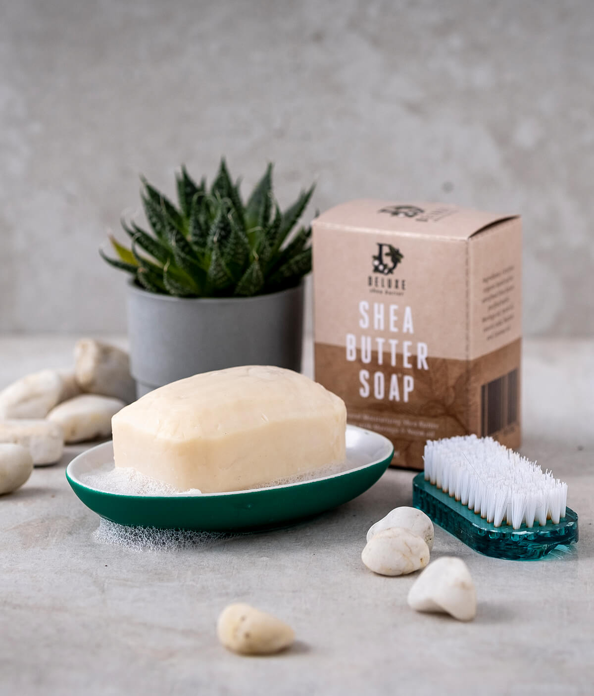Deluxe Shea Butter® Soap bar, in a dish with suds, next to product box and a scrubbing brush.
